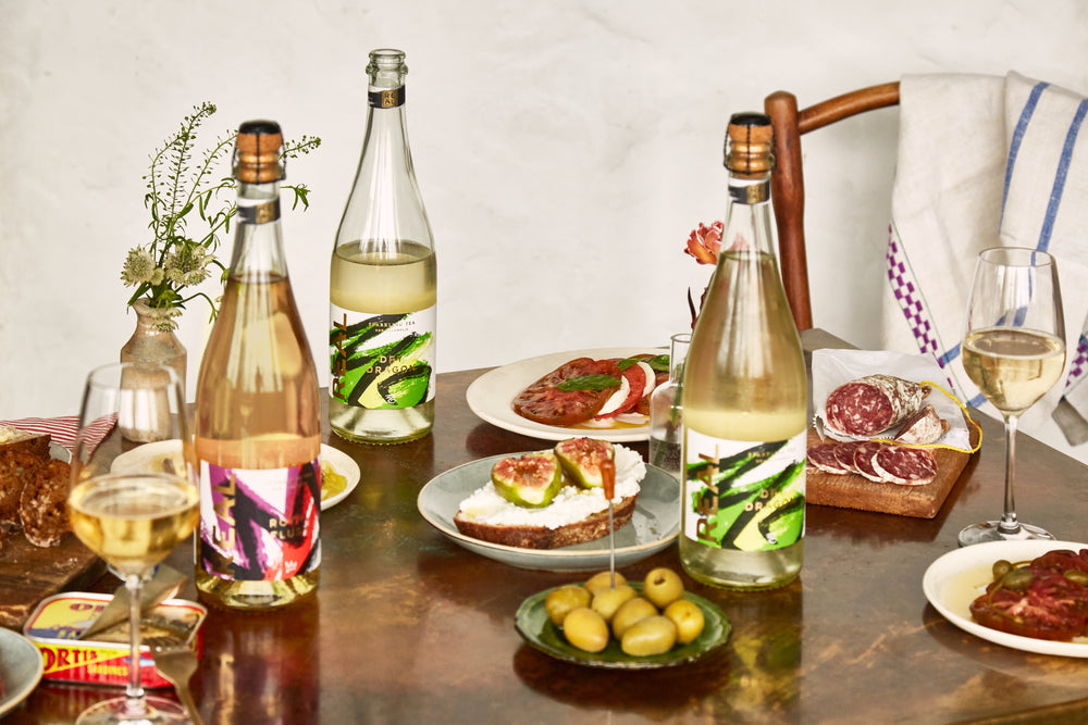 Bottles of the REAL Drinks Co Naturally Fermented Sparkling Tea sat on a table next to delicious foods, waiting to be indulged in 